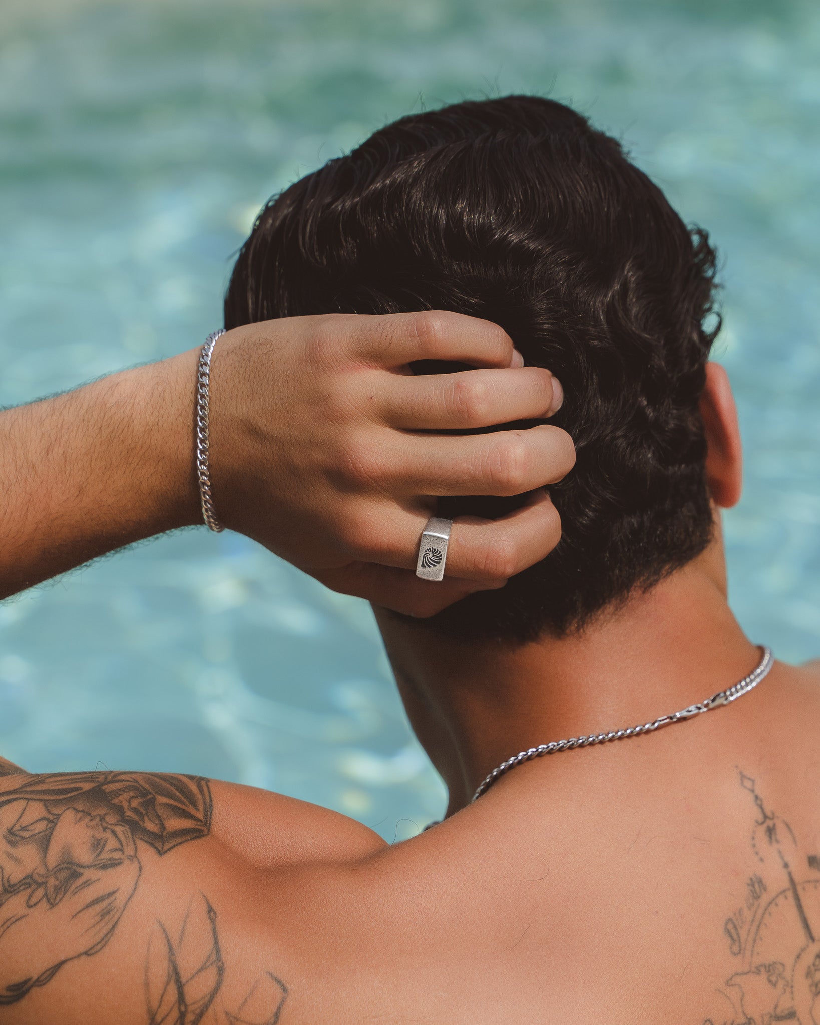 Vista lateral de un hombre junto a una piscina con anillo plateado cuadrado de concha en el dedo y pulsera plateada.