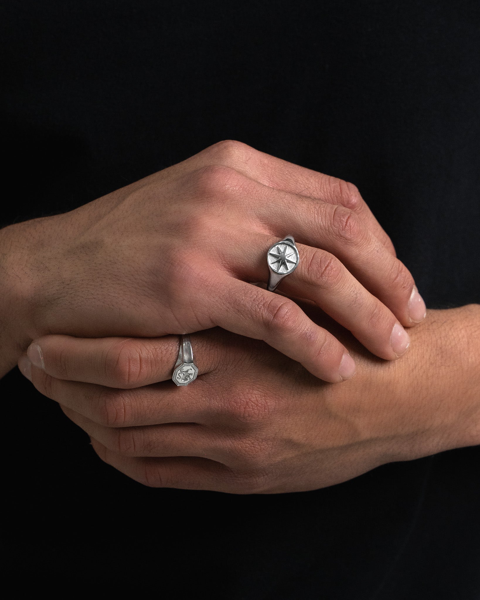 Dos anillos plateados en la mano, uno con escudo octogonal y otro con símbolo circular