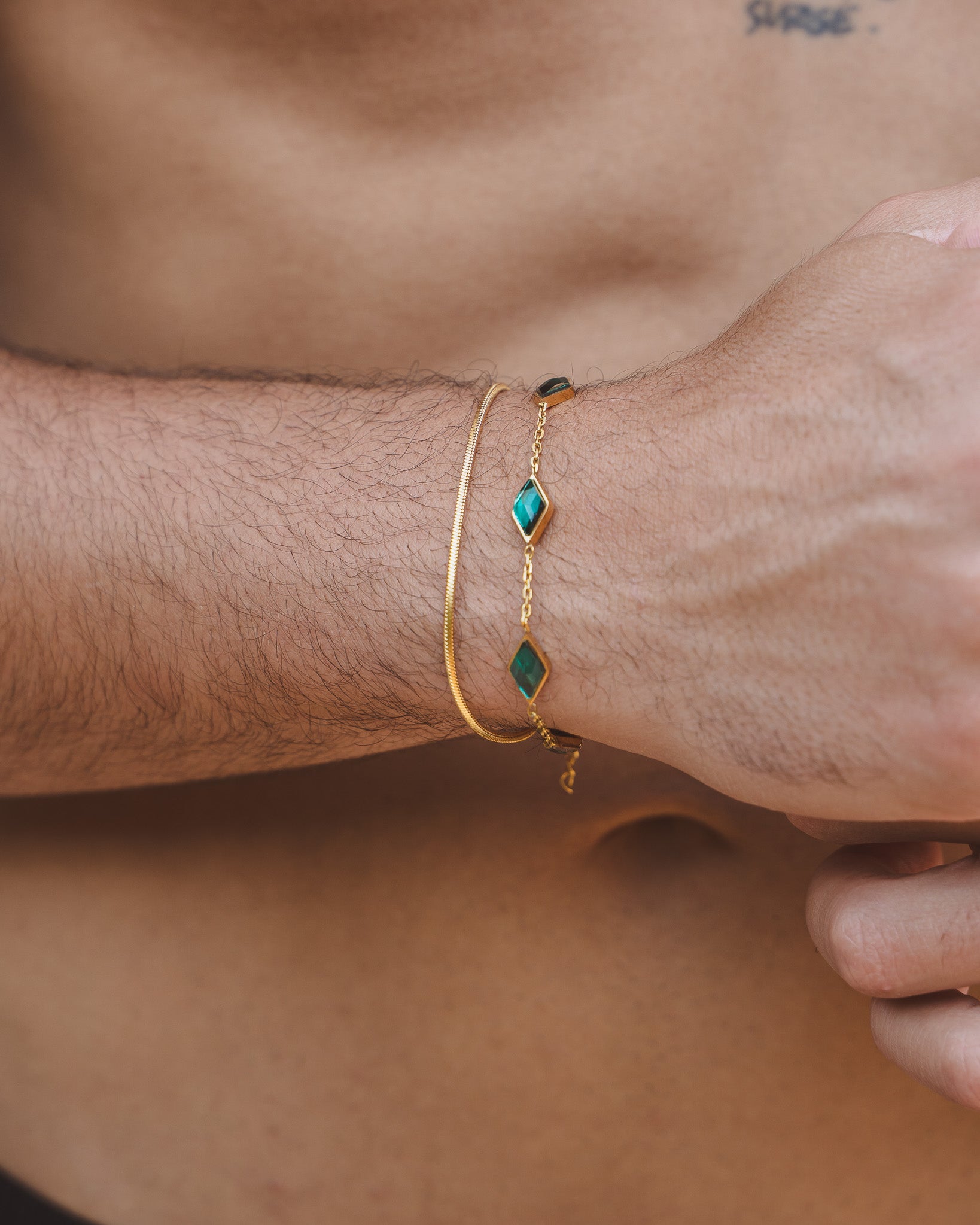 Amberes gold bracelet with emerald green diamond shaped stones on a wrist, paired with a slim gold bracelet.