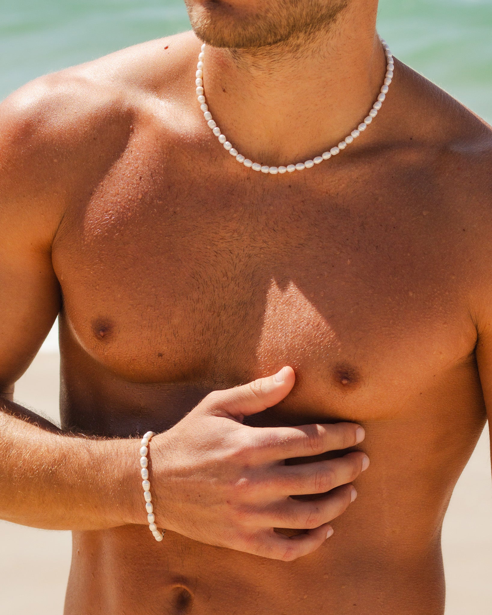 Shirtless man wearing Belize Pearl Chain white pearl necklace with visible tattoos on his arm at the beach