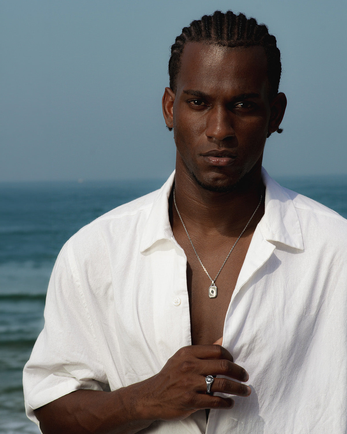 Man wearing a silver necklace with a textured rectangular pendant and a ring on his finger at the seaside.