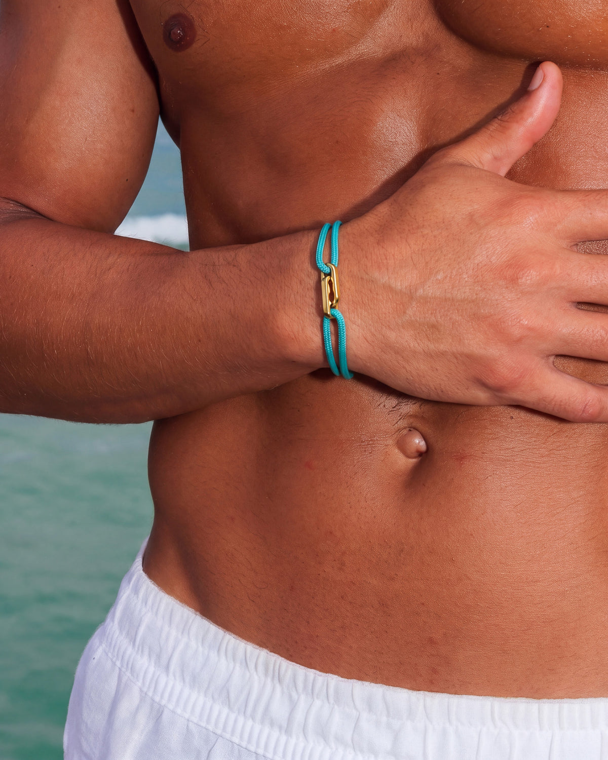 Pulsera dorada Atlantis con cordón turquesa en la muñeca de un hombre sin camiseta en la playa