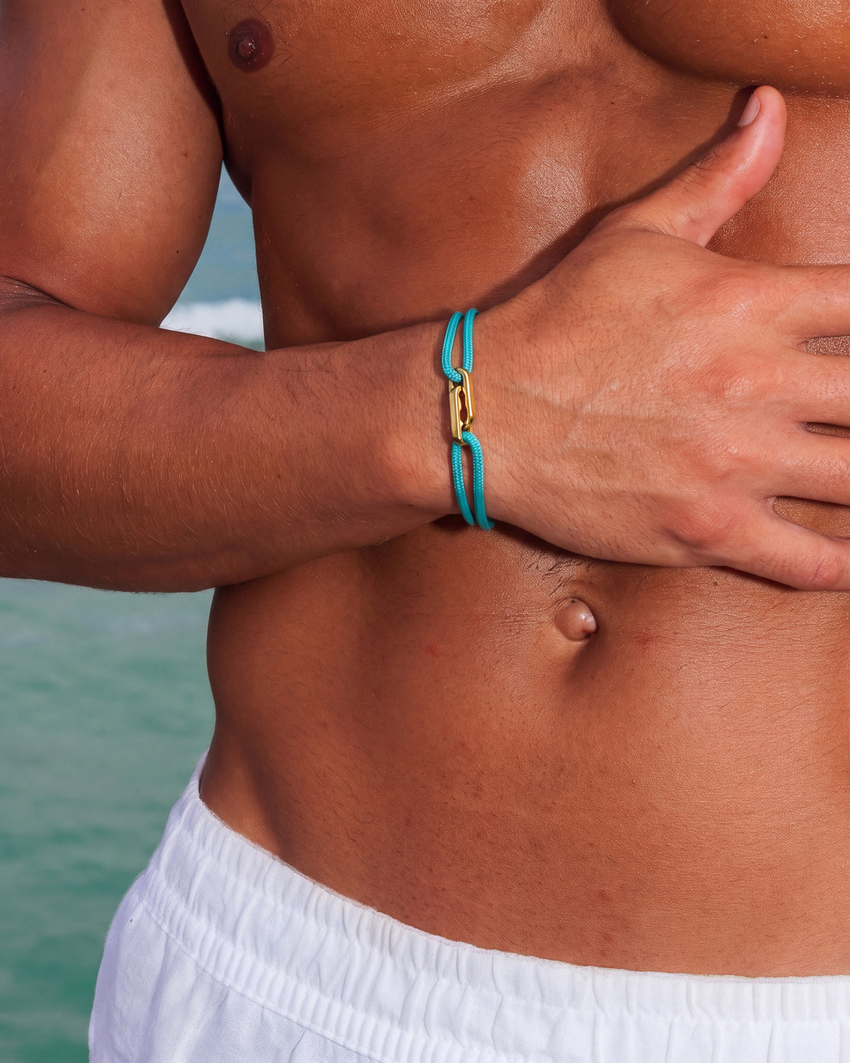 Pulsera dorada Atlantis con cordón turquesa en la muñeca de un hombre sin camiseta en la playa