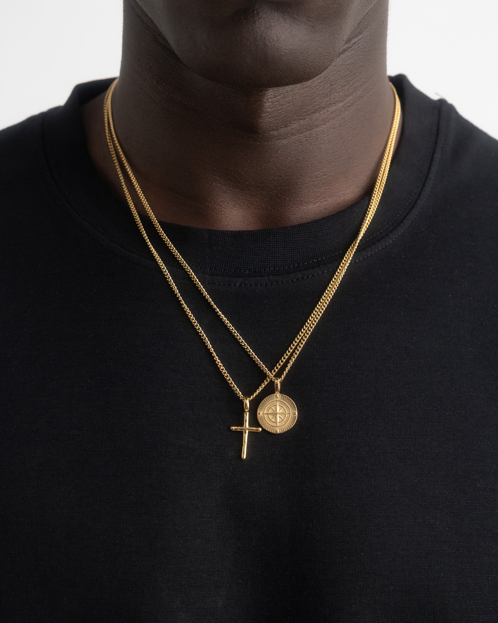 Close up of two gold chains with a round medallion and cross pendant on a dark shirt.