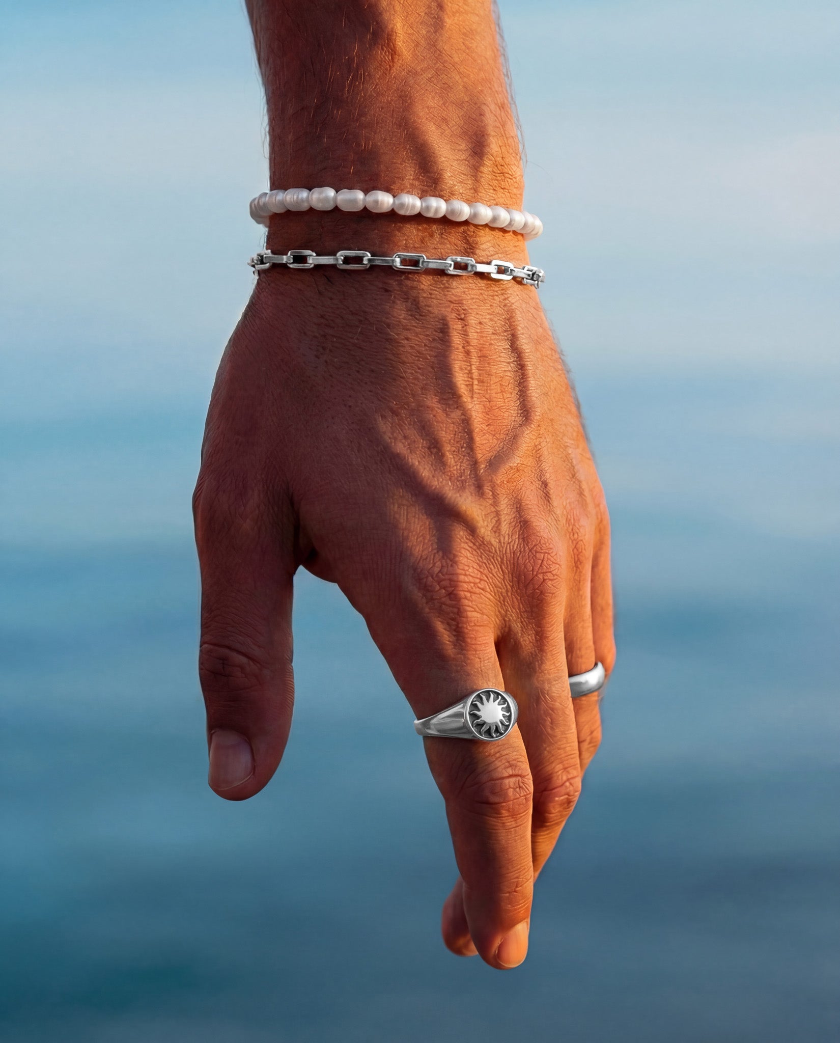 Daystar Silver Ring on a finger with pearl and silver chain bracelets against a blue sea backdrop.