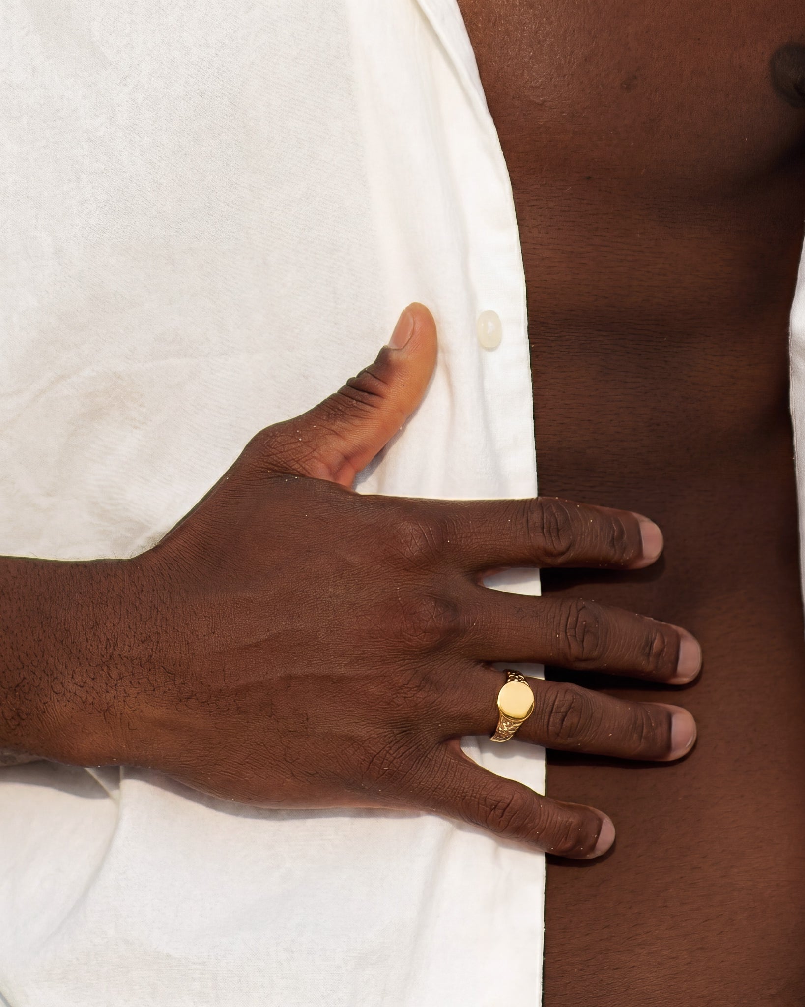 Gold Eden Ring with a smooth circular face on the ring finger of a dark-skinned hand resting on white fabric.