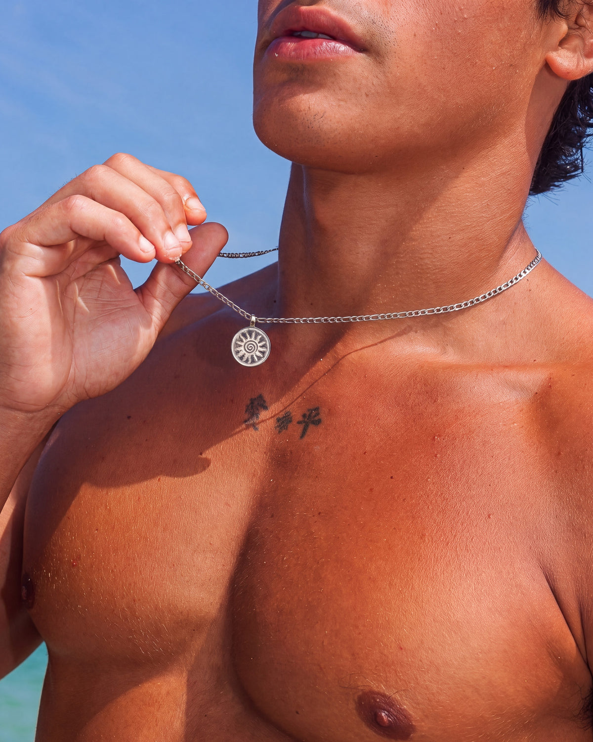 Primer plano del collar plateado Energy con colgante circular de sol sujeto entre los dedos contra un cielo azul.