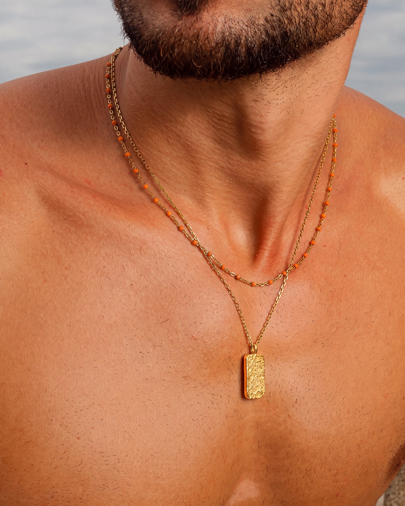 Bearded man wearing two gold chains with orange bead accents around his neck.