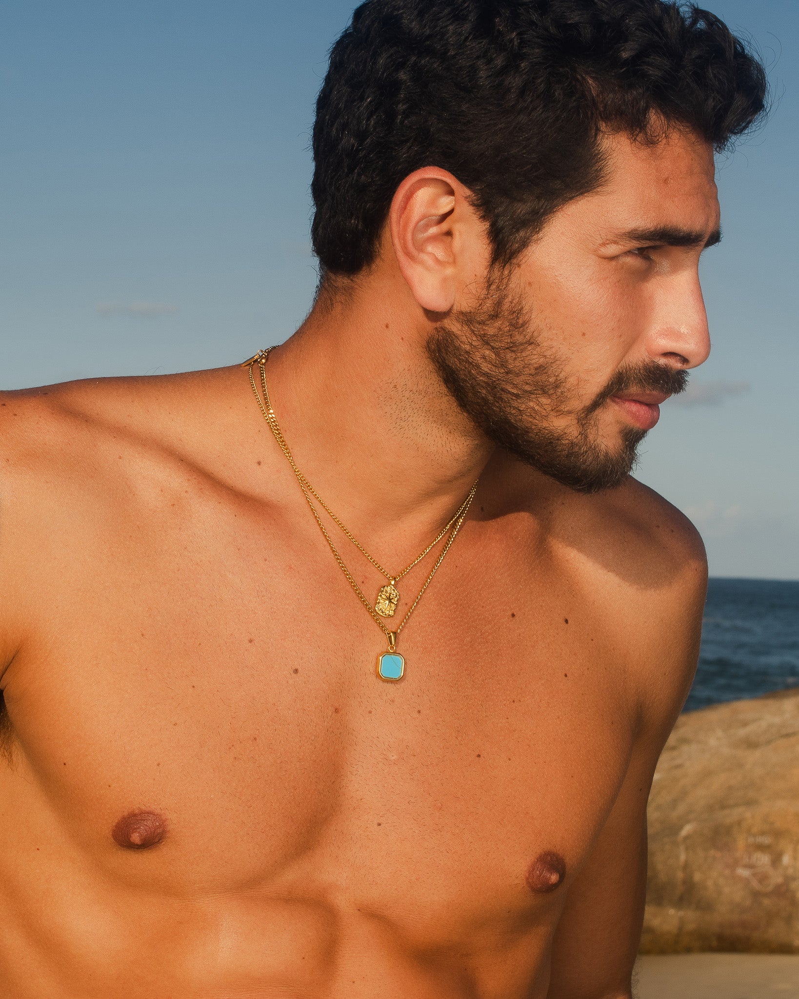 Primer plano de un hombre con collares dorados Nova en capas con colgante rectangular turquesa y pequeño charm dorado