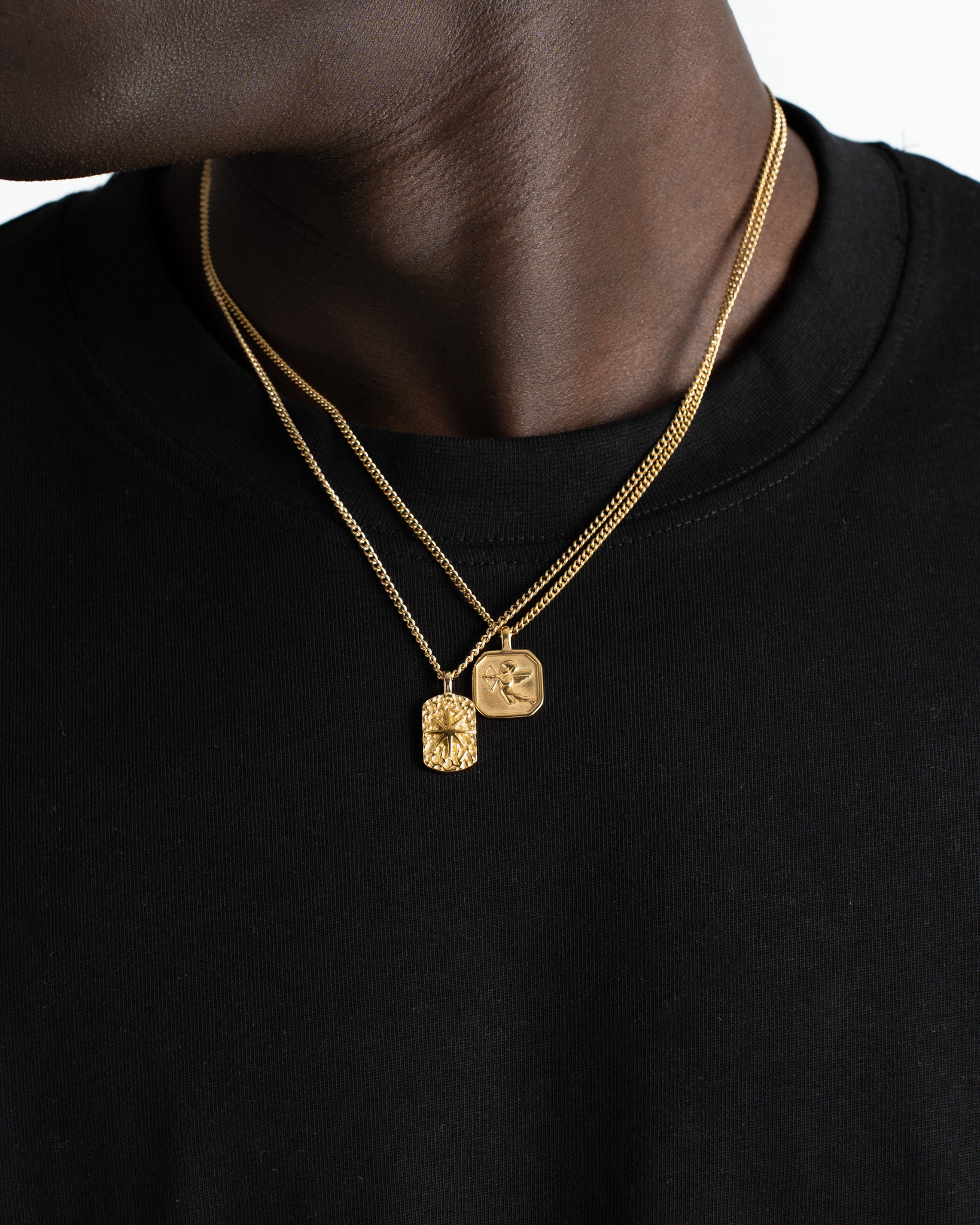 Gold chain necklace with two pendants a square engraved Cupid design and a textured rectangular charm on black fabric