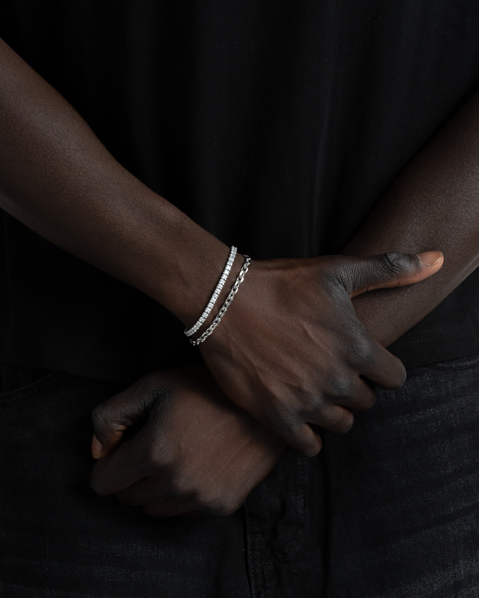 Wrist with a 4 mm icy tennis bracelet and a silver chain bracelet on a dark outfit