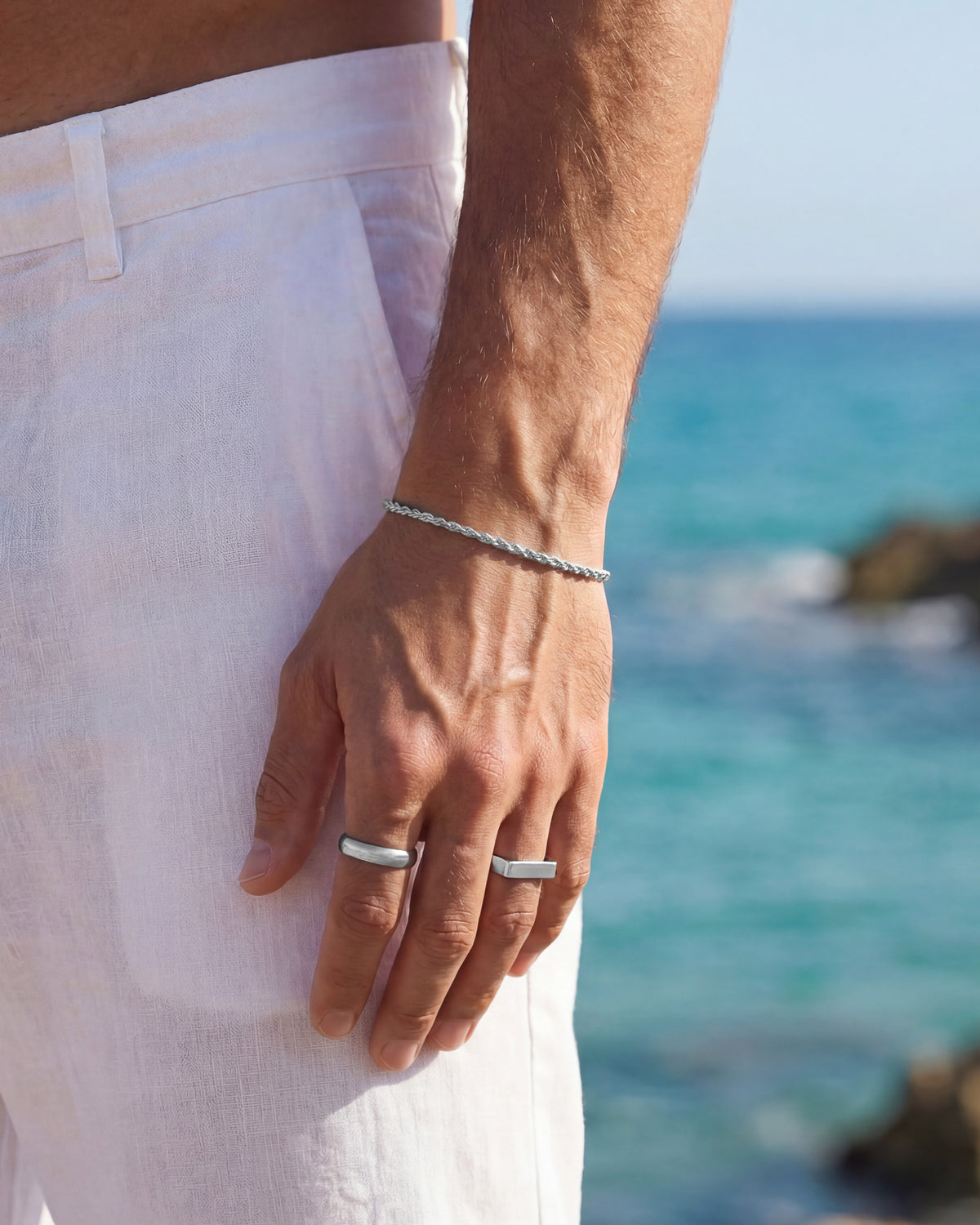 Pulsera plateada de cuerda trenzada en la muñeca de un hombre con pantalón corto blanco y fondo de mar.