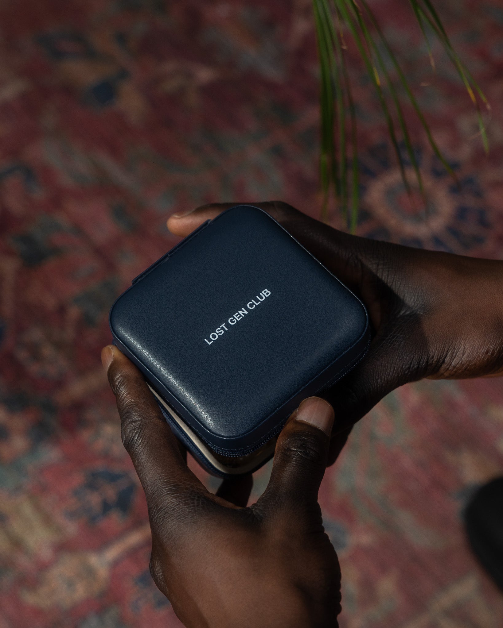 Close up of a dark blue travel jewelry case held by a hand against a colorful rug, logo visible on the lid