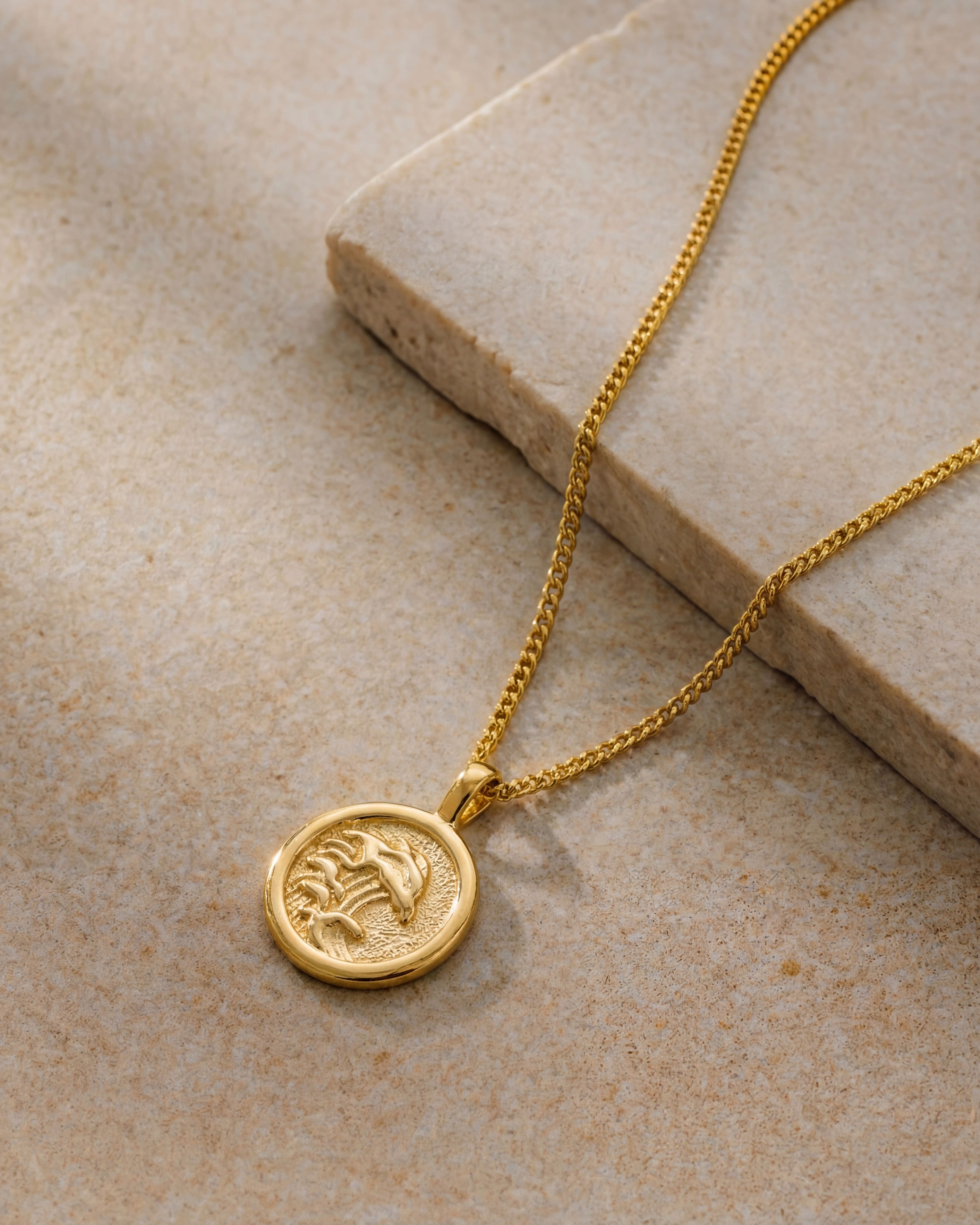 Close up of a gold palm tree pendant on a textured stone backdrop with chain