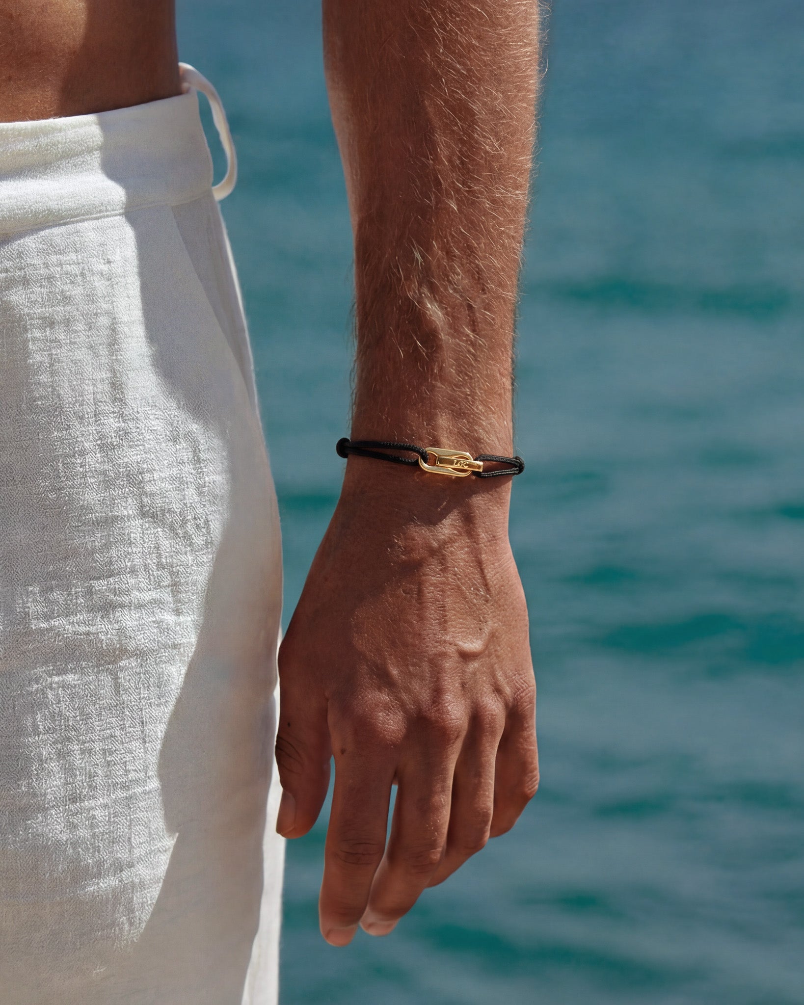 Atlantis gold bracelet with black cord and a gold clasp worn on the wrist by the sea