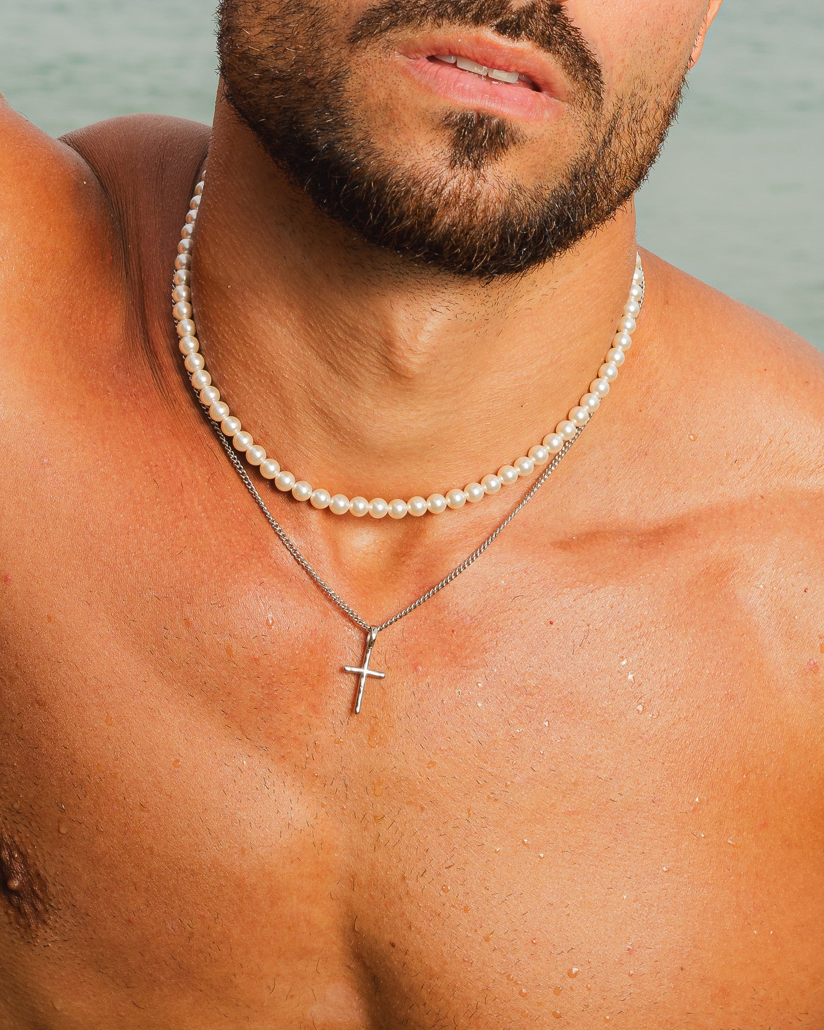 Silver cross necklace layered with a pearl strand on the chest.