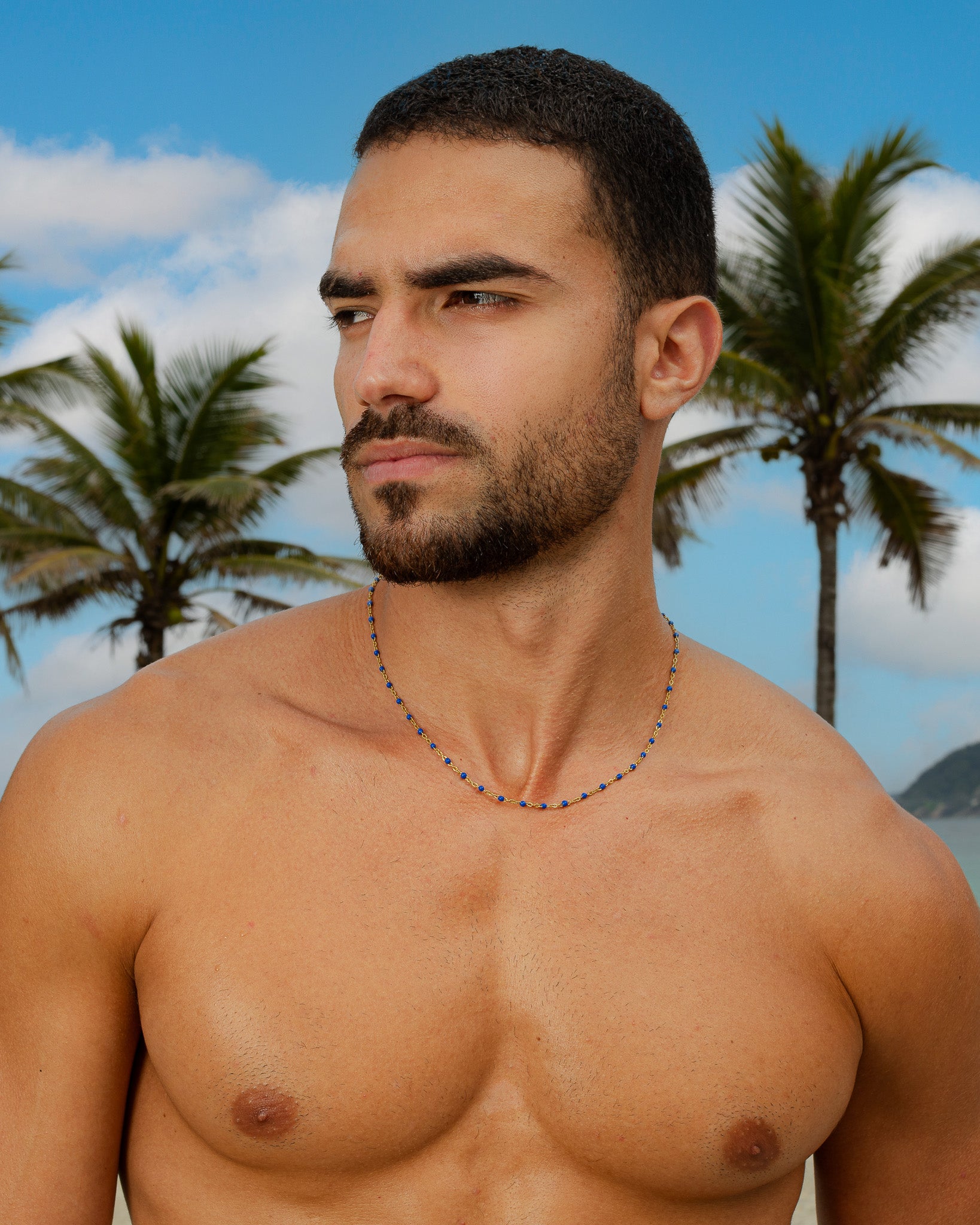 Man wearing a gold chain with blue beads against a tropical beach backdrop
