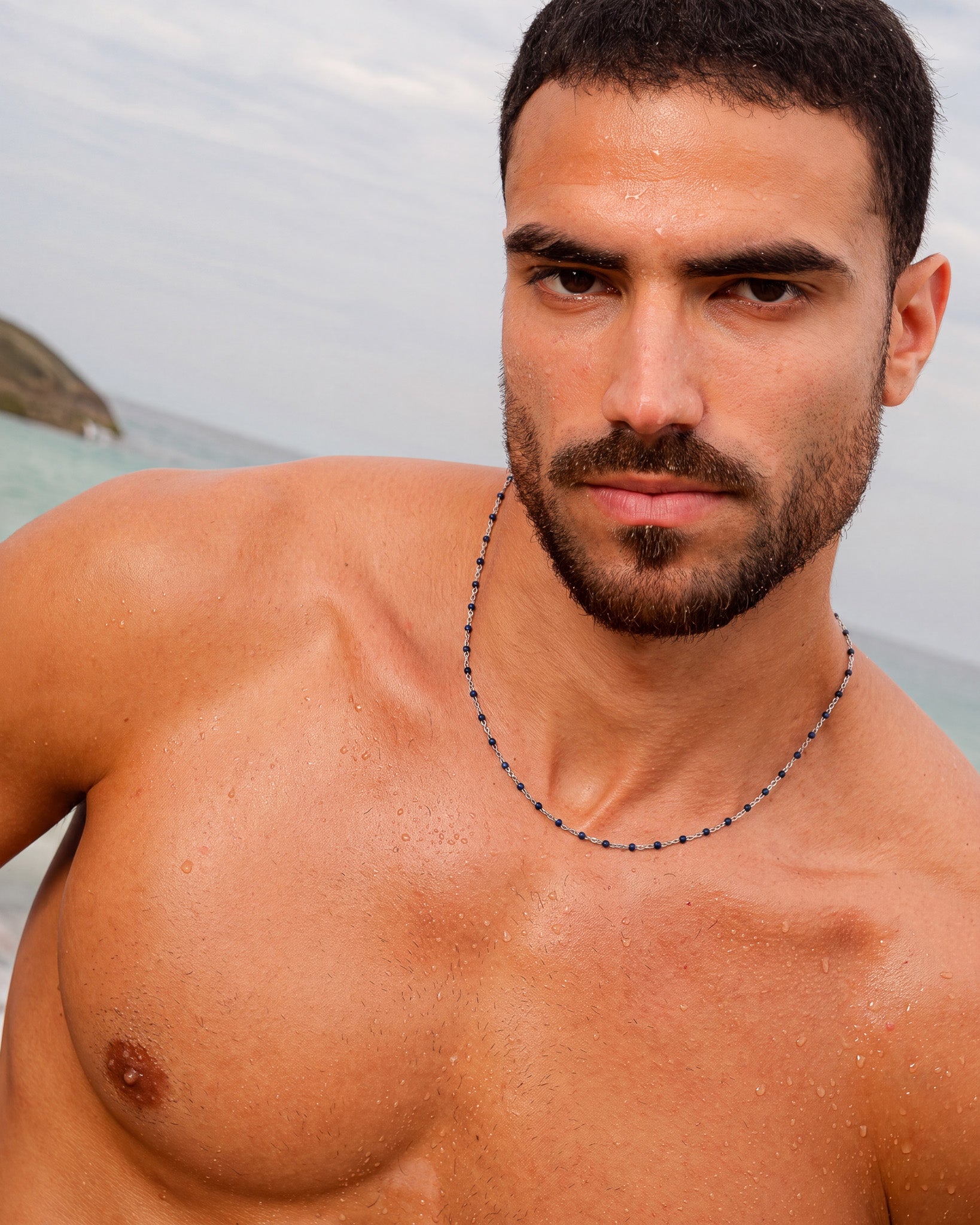 Silver chain necklace with blue beads worn by a shirtless man at the beach.