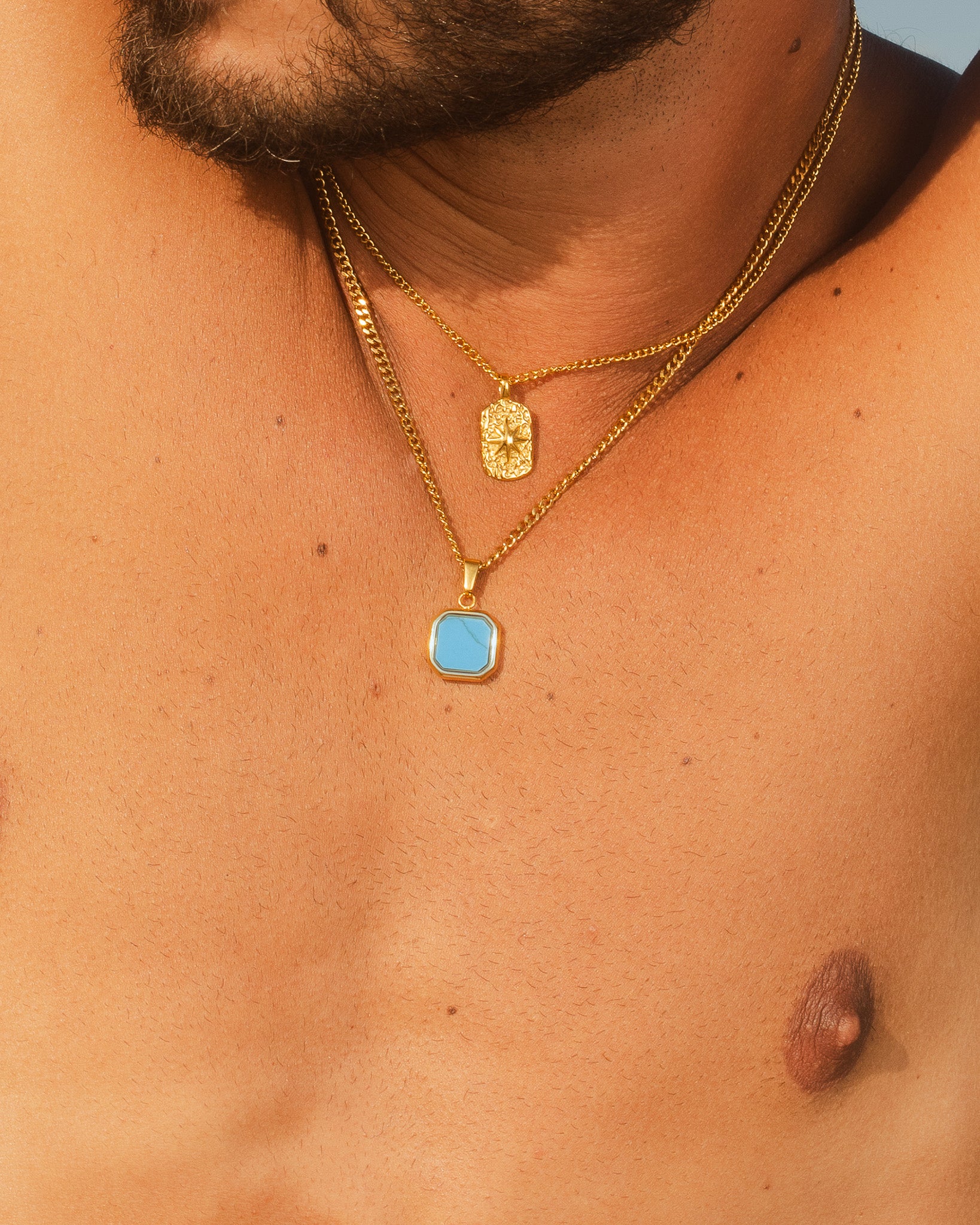 Chest shot of a man wearing multiple Nova Gold necklaces with turquoise hexagon pendant and two gold pendants against a rocky backdrop