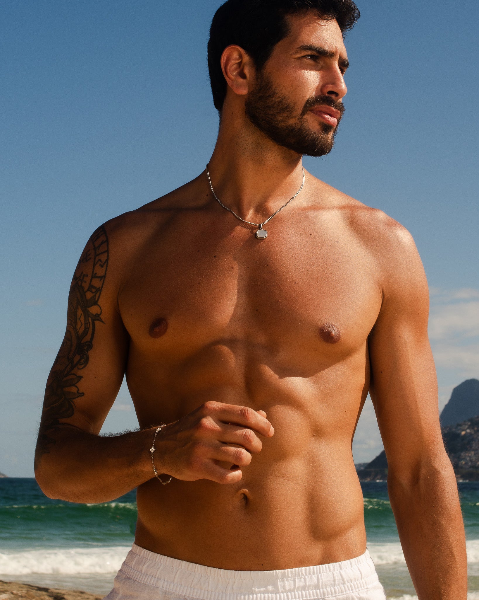 Clover silver bracelet on left wrist of a shirtless man at the beach